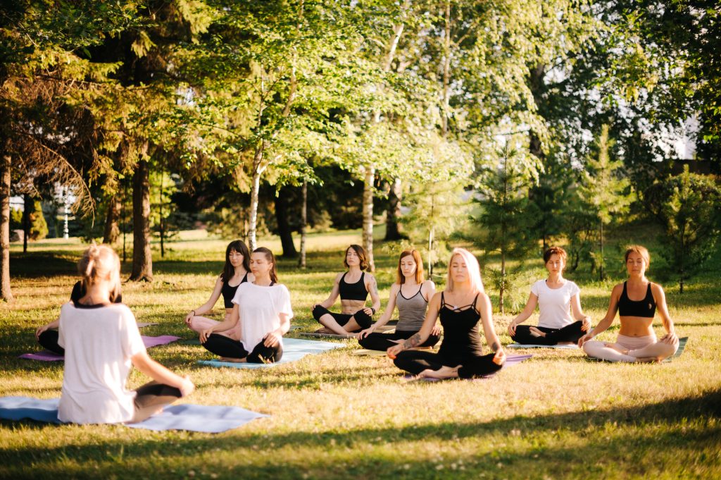 Group of young women are meditating in park on summer sunny morning under guidance of instructor. Group of girl outdoors are sitting in lotus pose on yoga mats on green grass with eyes closed